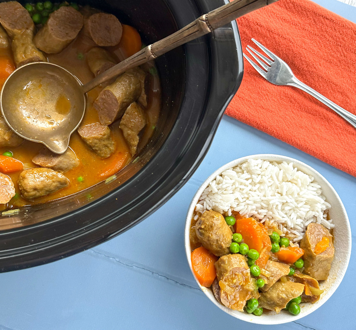Slow Cooker with curried sausages, and a white bowl with rice and curry sausages and a fork on a blue background