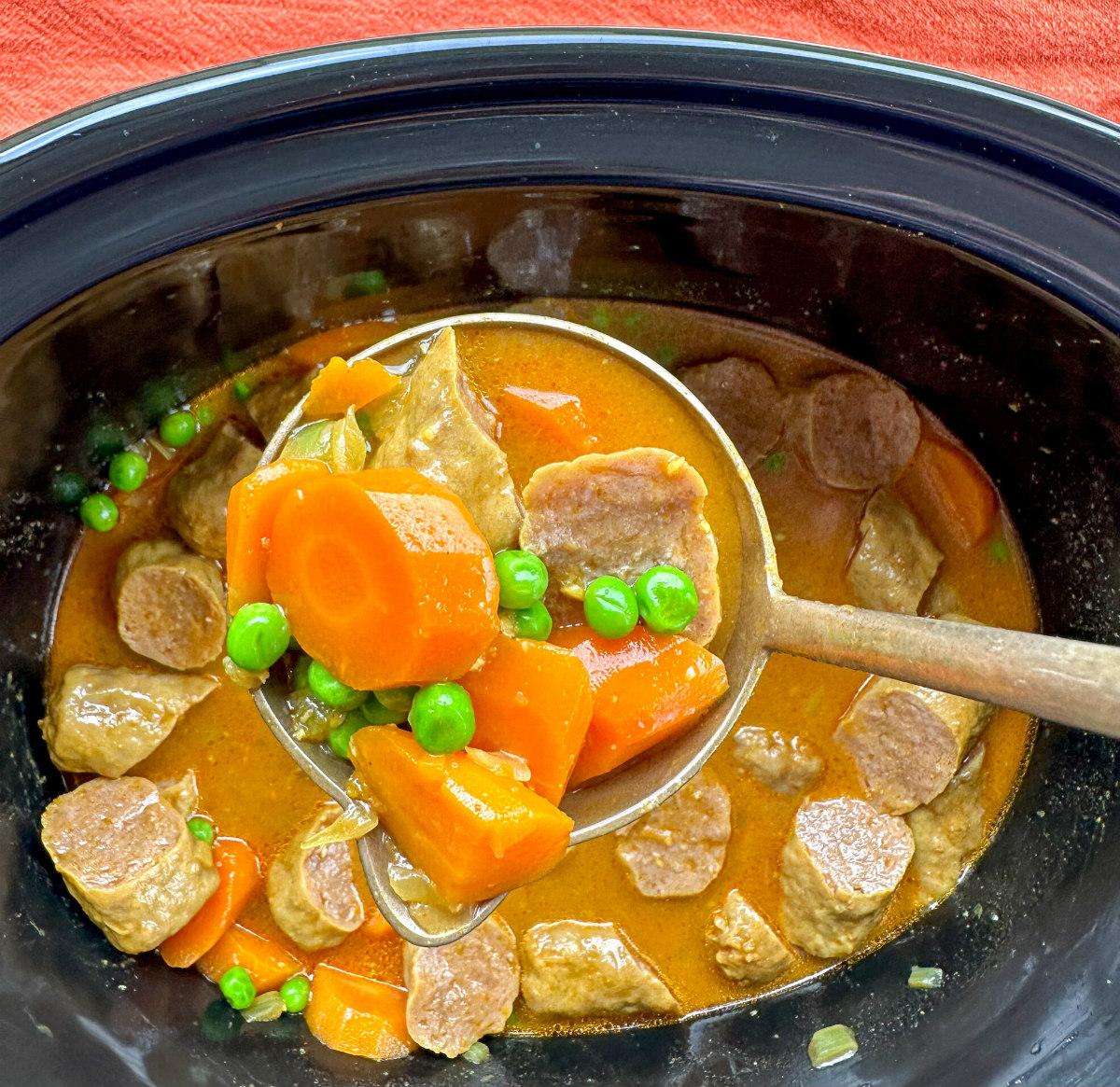 Slow cooker bowl with a ladle full of curried sausages, carrots and peas 