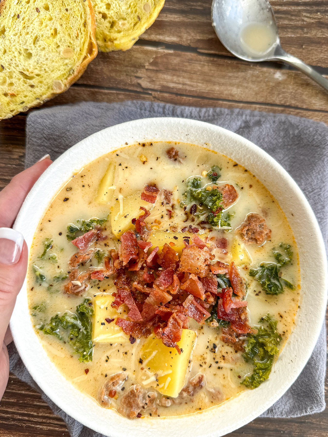 cozy bowl of Zuppa Toscana soup topped with crispy bacon and parsley with a hand holding it